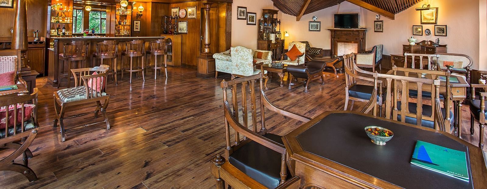 A bar and lounge area in naini retreat, nainital. The room features a wooden bar, comfortable seating arrangements, a fireplace, and a television. The overall ambiance is warm and inviting.