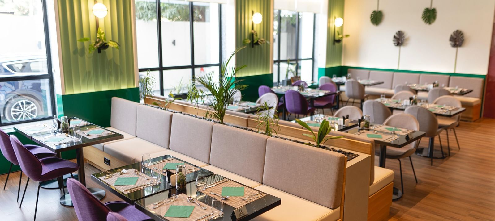 Bright, modern Meli-Melo restaurant interior with accents, large windows, potted plants, and neatly set tables with chairs and beige booth seating. Natural light fills the stylish dining area