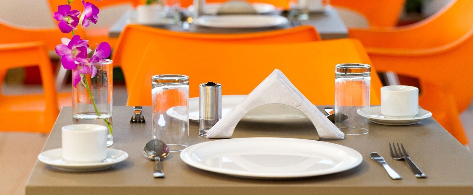 A neatly set outdoor dining area at Sanctum Suites Whitefield, Bangalore featuring tables and bright orange chairs with white tableware.