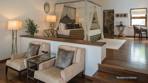 A bedroom at The Serai Bandipur with a four-poster bed, chairs, side tables, lamps, artwork, and wooden flooring, divided by a small steps.