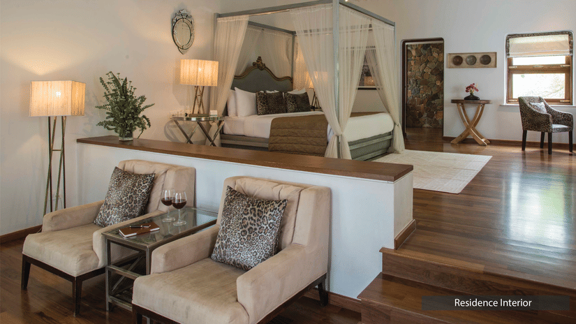 A bedroom at The Serai Bandipur with a four-poster bed, chairs, side tables, lamps, artwork, and wooden flooring, divided by a small steps.
