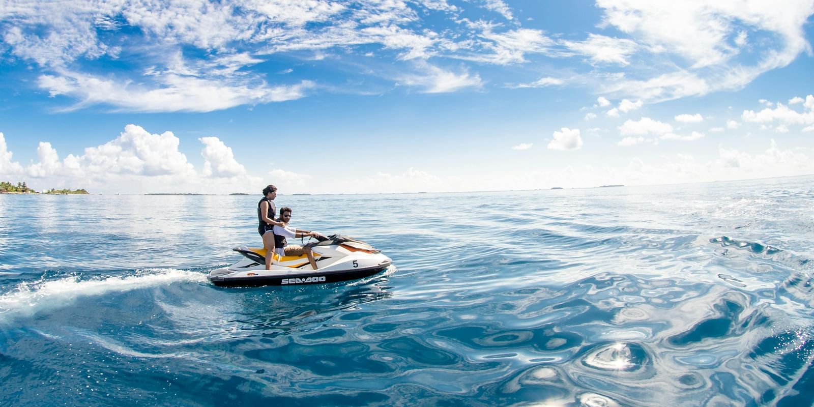 A couple in a motor boat cruising through the lake. At the horizon there are some trees and up above there is a bright sky.