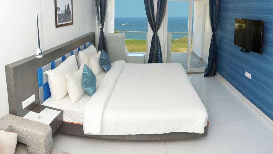 A hotel bedroom with a large white bed, blue accent wall, and a view of the outdoors through a large window | Grande Chalet Sea View | Grande Bay Resort & Spa, Mamallapuram