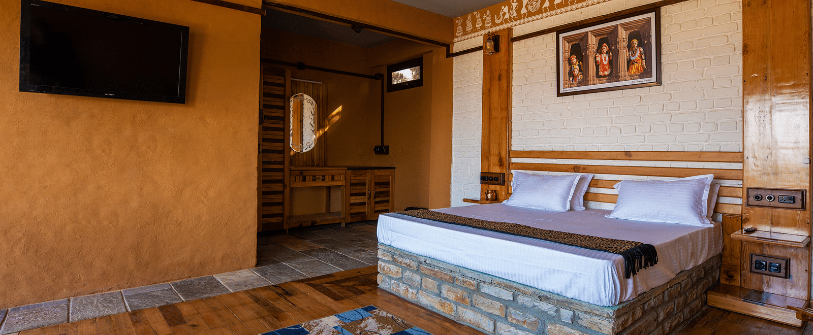 Rustic hotel room with wooden bed, wall art, TV, and patterned rug on hardwood floor. - Blusalzz Homes The Himalayan Bungalow, Almora