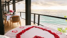 An outdoor terrace with a bed decorated with a heart made of rose petals overlooks a scenic ocean view.