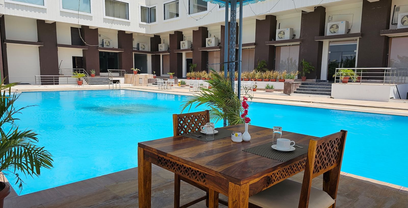 Shot of an outdoor dining area at the Bamboo Saa Mulberry Resort, Pushkar, featuring a wooden table and two chairs set beside a bright blue swimming pool.