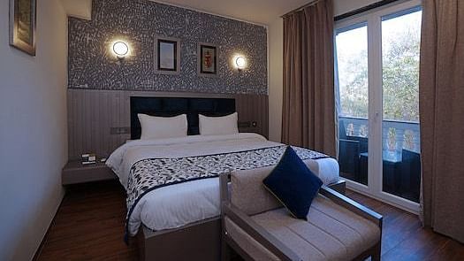 A hotel bedroom at Grand Continent, Anjuna featuring a large bed, a sofa at the foot, and a balcony with open curtains visible on the right. The wall behind the bed has decorative textured panelling and sconce lights.