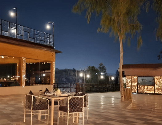 An outdoor dining area at night with a table and chairs set on a paved patio. Lights illuminate the space.- Bamboo Saa Resort & Spa, Udaipur.
