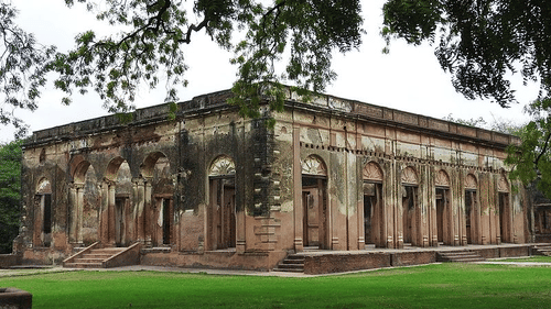 A low, historical building with numerous arched windows sits on a large, well-kept green lawn surrounded by trees.