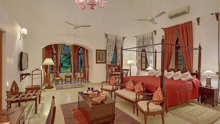A well-appointed room with a four-poster bed, seating area, and decorative rugs, featuring arched windows and a chandelier- Shahpura Bagh.