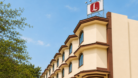 Exterior view of a multi-story cream-coloured hotel building with a red rooftop Lords logo sign, paved driveway, and a parked silver car in front - Lords Inn Vadodara