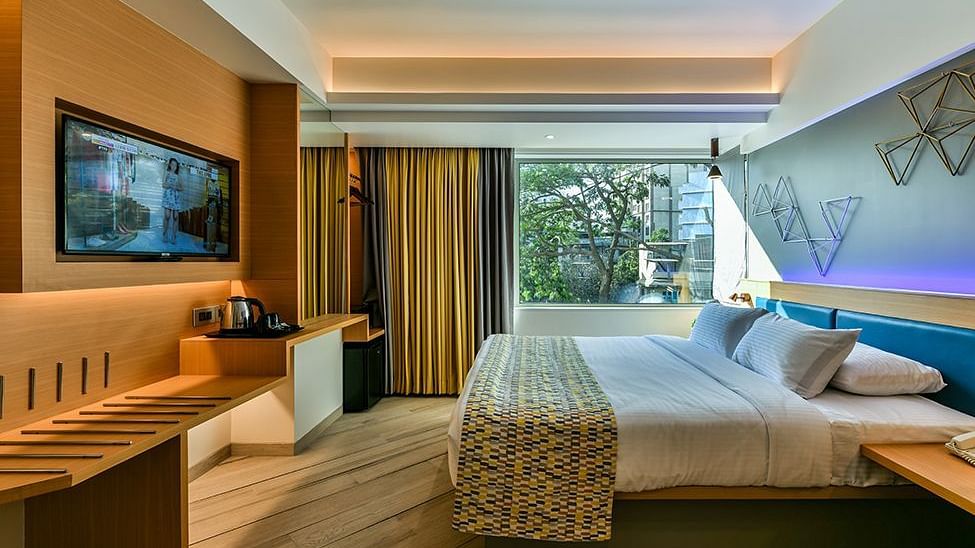 A spacious room with a large bed, wooden bedside table, a wall-mounted television opposite the bed with a wooden table beneath it, and the window curtains pulled to the side at Indie Stays