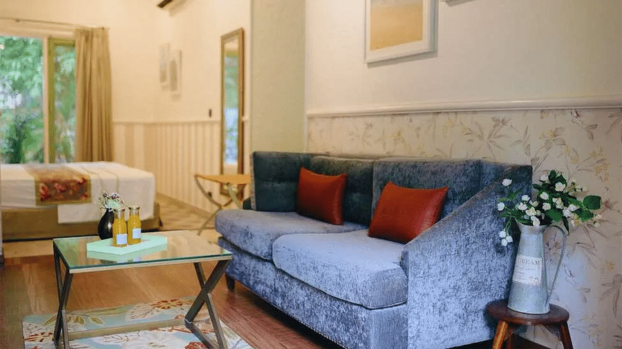 Interior of Karma Chalets with a grey sofa, center table, cushions, rug, artwork, and a window with greenery outside.