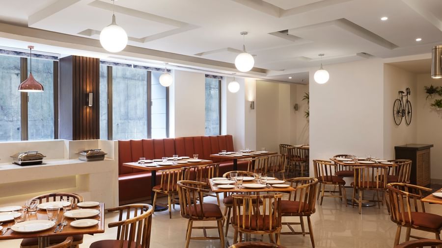 A spacious, modern dining hall at Monday Hotels Mahape, Navi Mumbai, with multiple wooden tables, chairs, and a long booth.