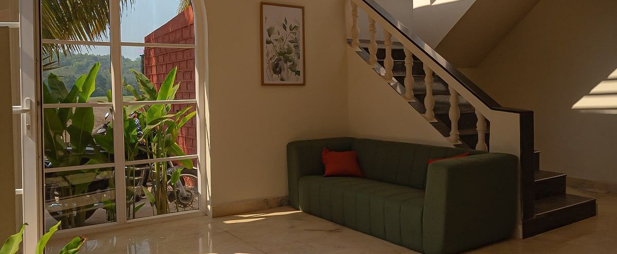 An interior view looking up a curved staircase with wooden treads and a white balustrade. Sunlight streams in from above, illuminating a seating area with a dark sofa and plants on the ground floor at Kssemya
