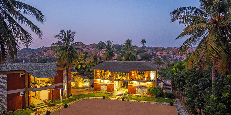 The resort buildings illuminated at dusk, showcasing the red-brick architecture and sandy courtyard - Nature Trails Ashoka Resort Hampi