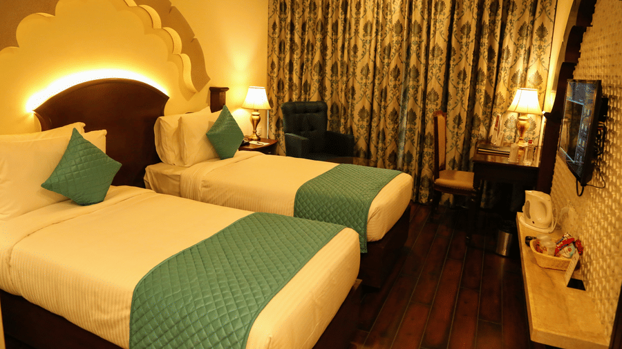 A bedroom at Comfort Inn Sapphire by Eastlynn Collections, Jaipur with 2 single beds, headboard arch, curtains, and side table with lamp.