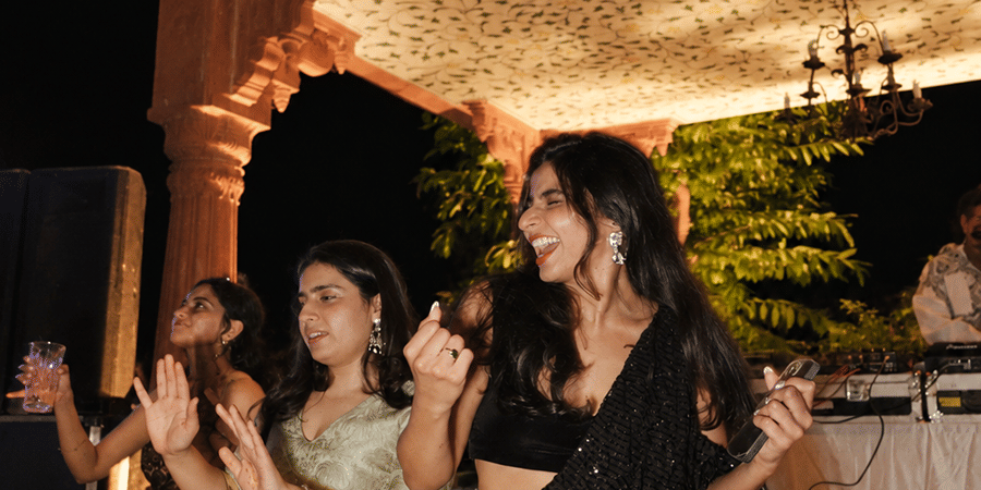 Two women happily dancing together during an evening event on a lighted outdoor pool side at Umaid Palace.