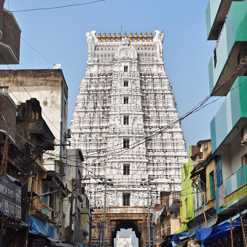 A view from afar of the majestic Govindaraja Temple in Tirupati, with people walking in front of it on the road, as seen during the day.