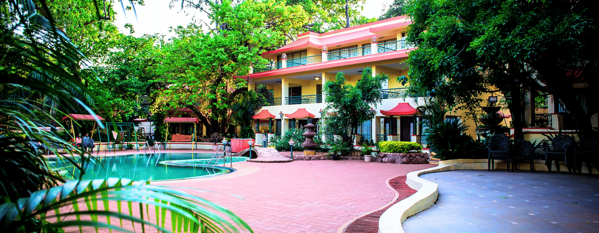 A partial view of the pool with its front pathway and the hotel building behind while being surrounded by the trees of the property.