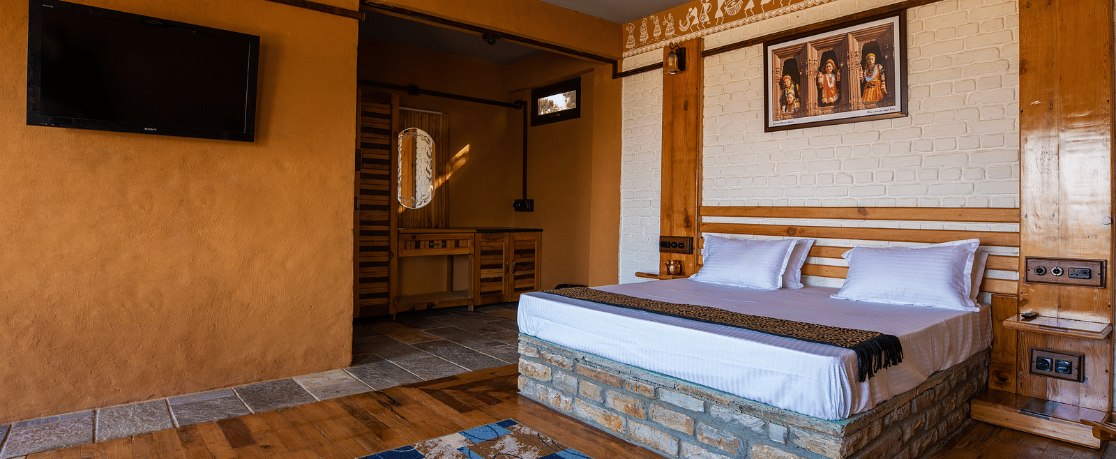 Rustic hotel room with wooden bed, wall art, TV, and patterned rug on hardwood floor. - Blusalzz Homes The Himalayan Bungalow, Almora