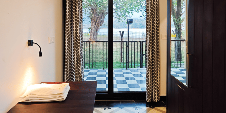 View from inside a guest room looking through glass sliding doors toward a tiled balcony and lush greenery - Ibex Resorts, Malampuzha (Kava Eco Camp and Caravan Park)
