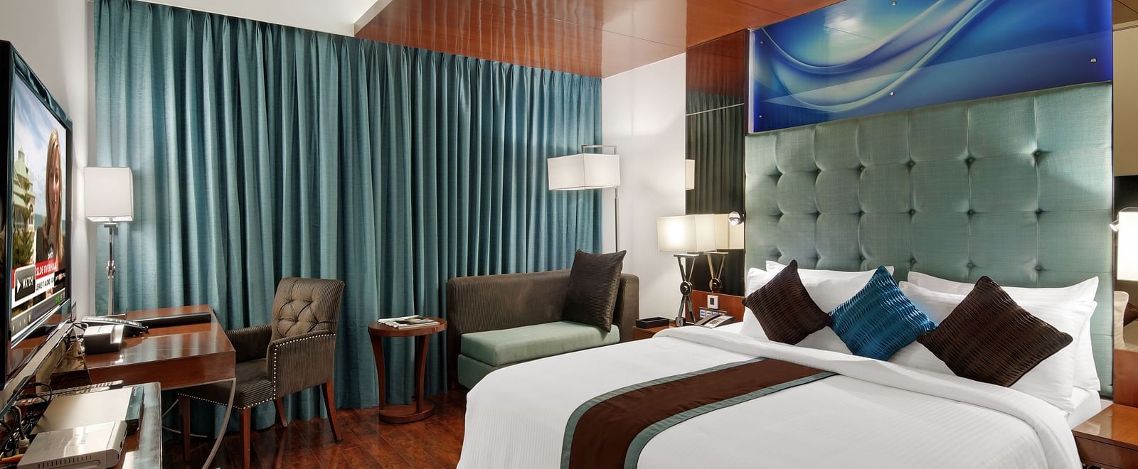 A spacious deluxe hotel room with a large double bed, decorative cushions, wooden flooring, a padded headboard, a wooden desk with chair, soft lighting, and curtains