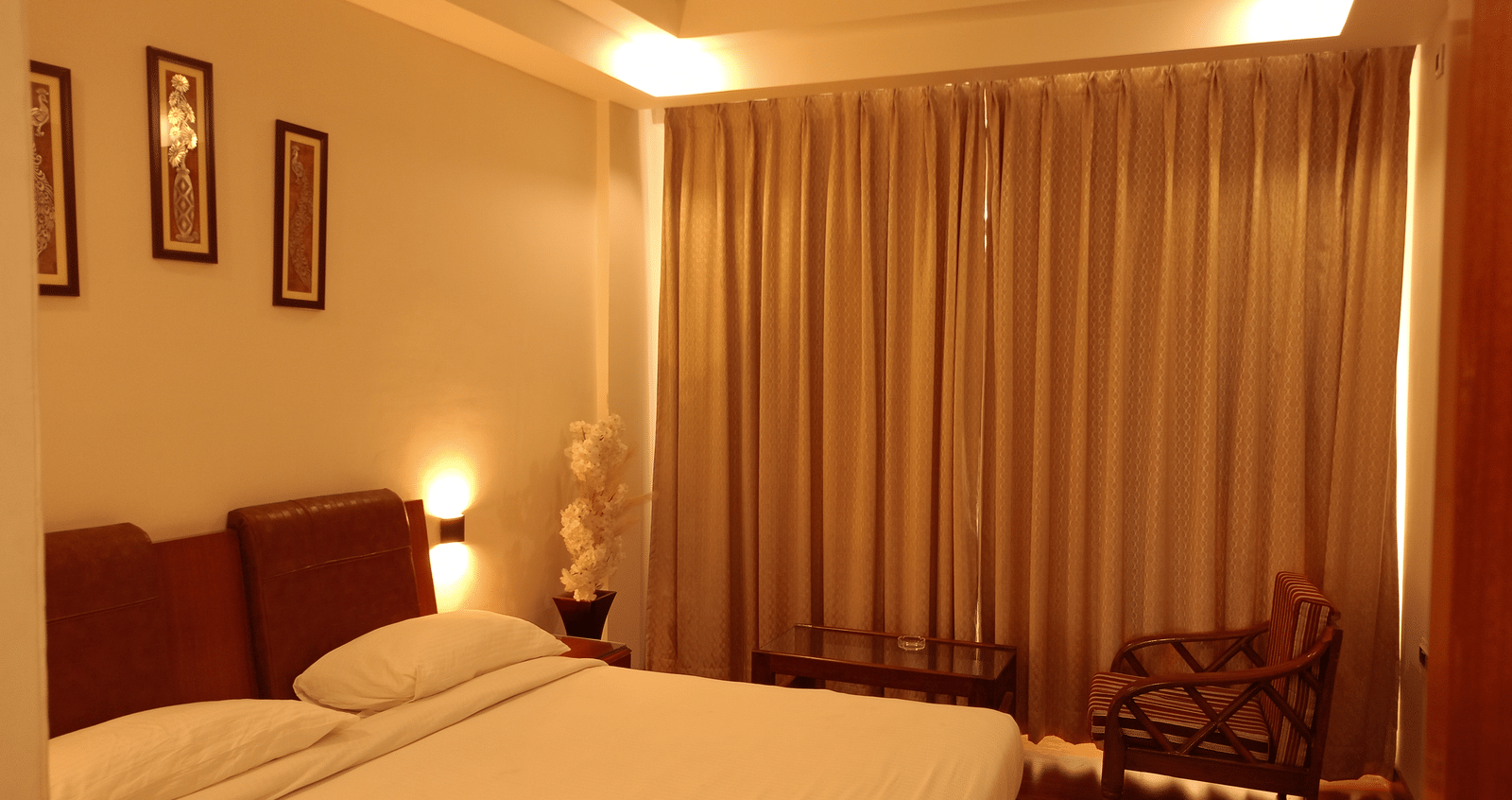 Guest room at Hotel Gold, Panipat featuring a bed, chair, side tables, and large window curtains.