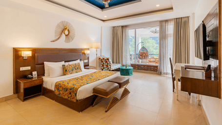 A well-lit bedroom featuring a big bed facing a television screen, with an attached balcony featuring a standing swing chair overlooking the trees outside | Summit by the Ganges Beach Resort & Spa, Rishikesh