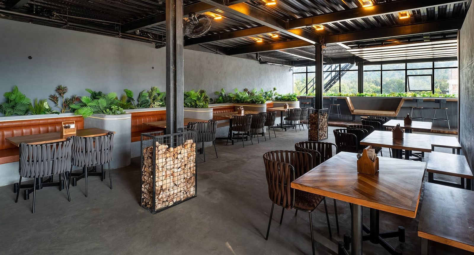 A modern open-air restaurant with wooden tables, cushioned seating, industrial-style ceiling, and lush greenery, offering a stylish dining experience at TGI Livancia Hillside, JC Nagar, Mysore.