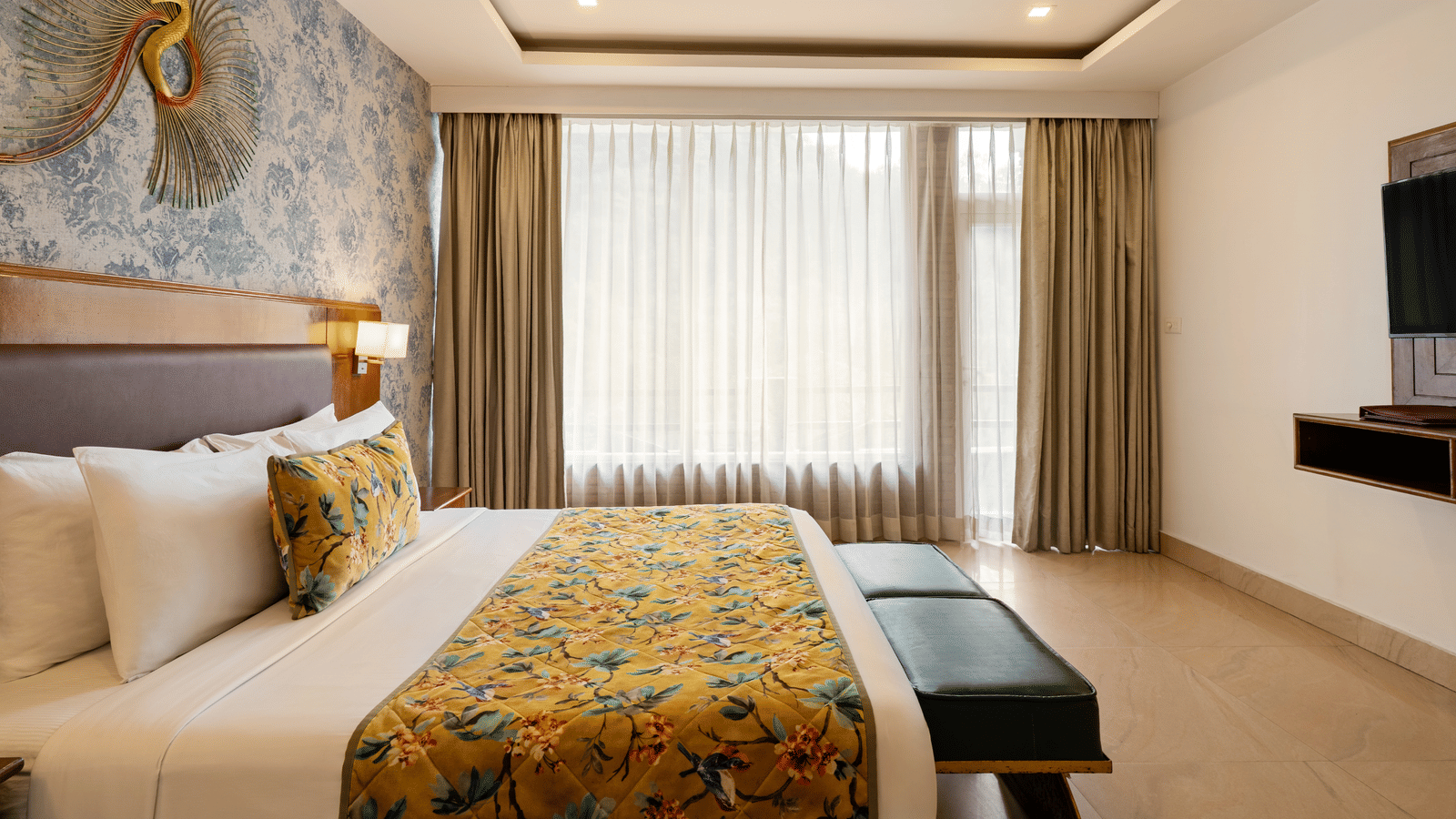 A bedroom with a neatly made bed facing a television unit, a decorative art piece over blue printed wallpaper, and full-length curtains | Summit by the Ganges Beach Resort & Spa, Rishikesh