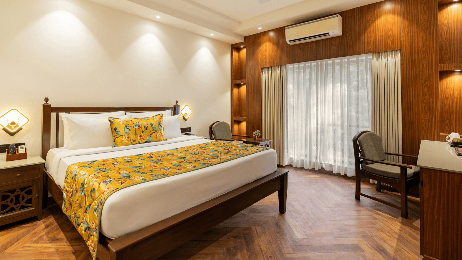 A spacious bedroom with an attached balcony, featuring a big bed, 2 bedside tables, 2 wooden chairs, a small table with a plant, an A/C unit and a fan, and a wooden floor | Summit by the Ganges Beach Resort & Spa, Rishikesh
