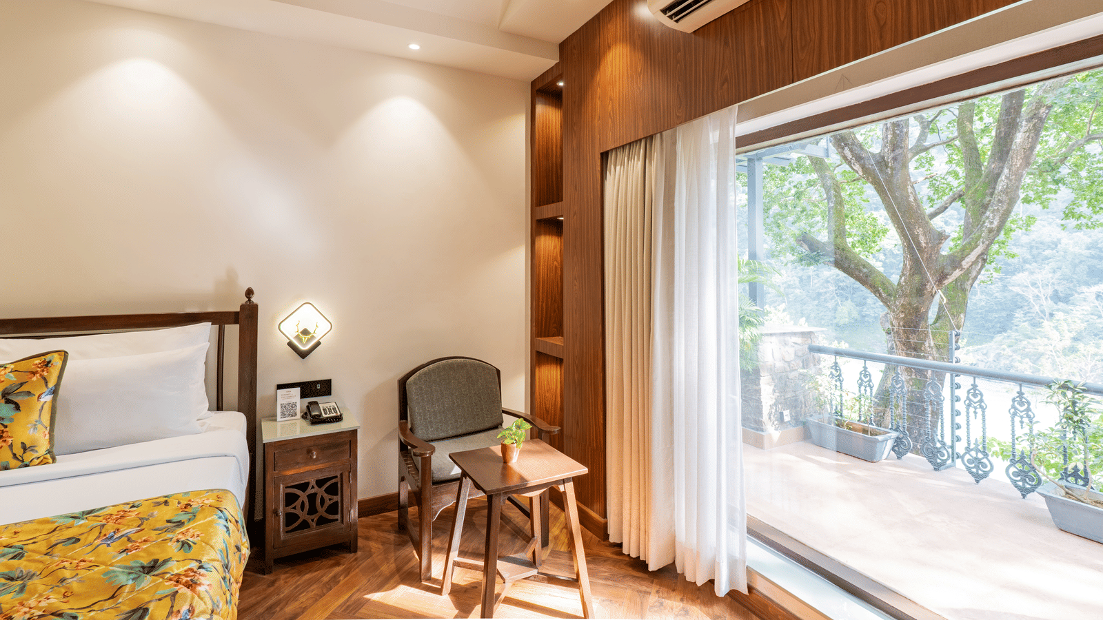 A bedroom with an attached balcony that has potted plants, decorative metal railing, a tree outside, and view of the river and trees in the distance | Summit by the Ganges Beach Resort & Spa, Rishikesh
