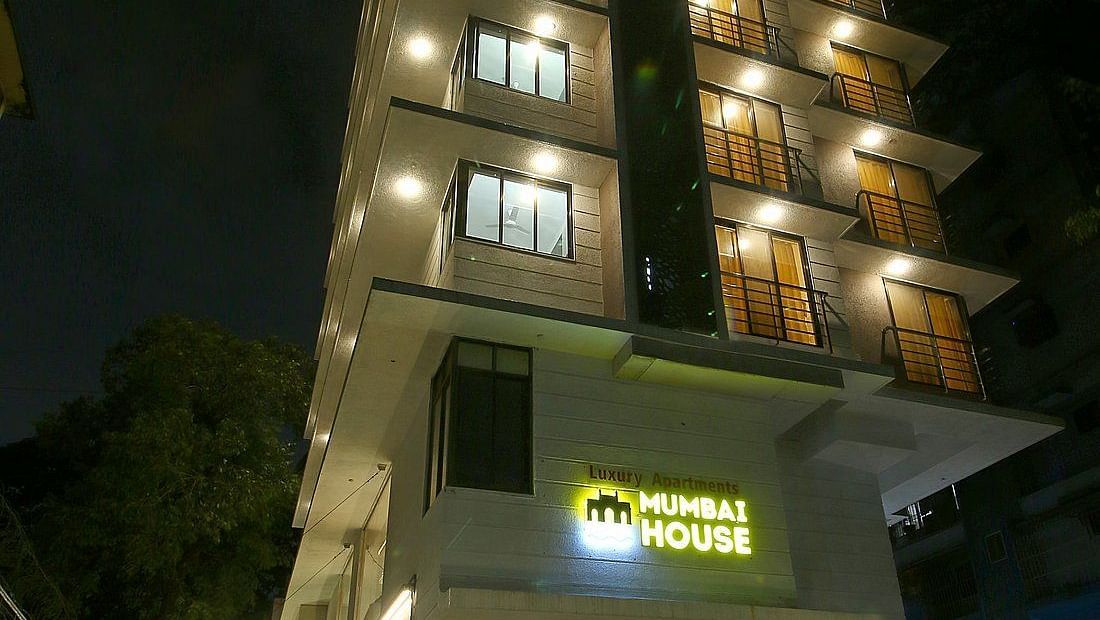 The exterior of Mumbai House at night, showcasing its multi-story structure with lit windows and a sign indicating it's a luxury apartment building.