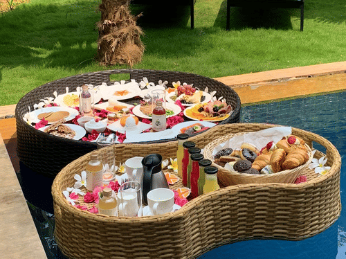 A close up of Two baskets filled with food, placed on the pool with blue loungers kept on the lawn area next to the pool at Azaya Beach Resort, Goa.
