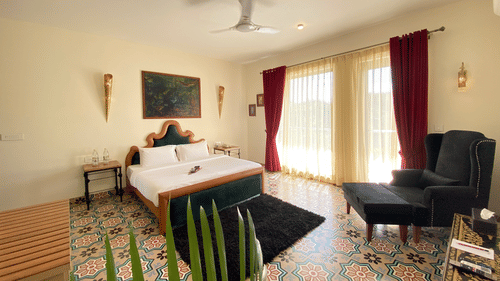 A bedroom with patterned floor tiles, a bed with a headboard, and an armchair. A ceiling fan, two wall sconces, a painting, two bedside tables, and large glass windows with curtains - Karma Lakelands