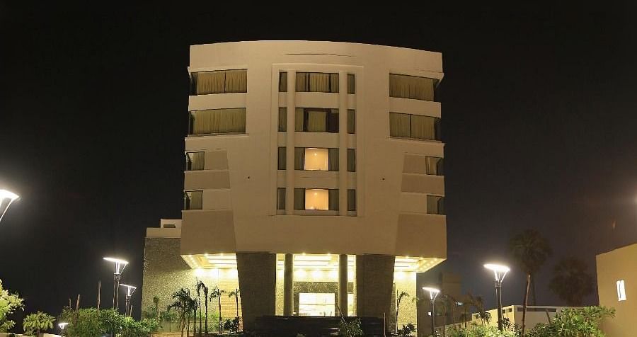 The exterior of Dhanalakshmi Srinivasan Hotels, Perambalur, a multi-story building with light-coloured facades and yellow-tinted windows, seen from a paved road under a blue sky.