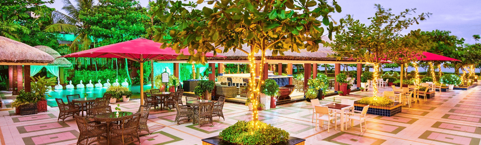 Large outdoor patio or courtyard area with green and pink lighting, an ornamental planter in the center, and scattered tables and chairs