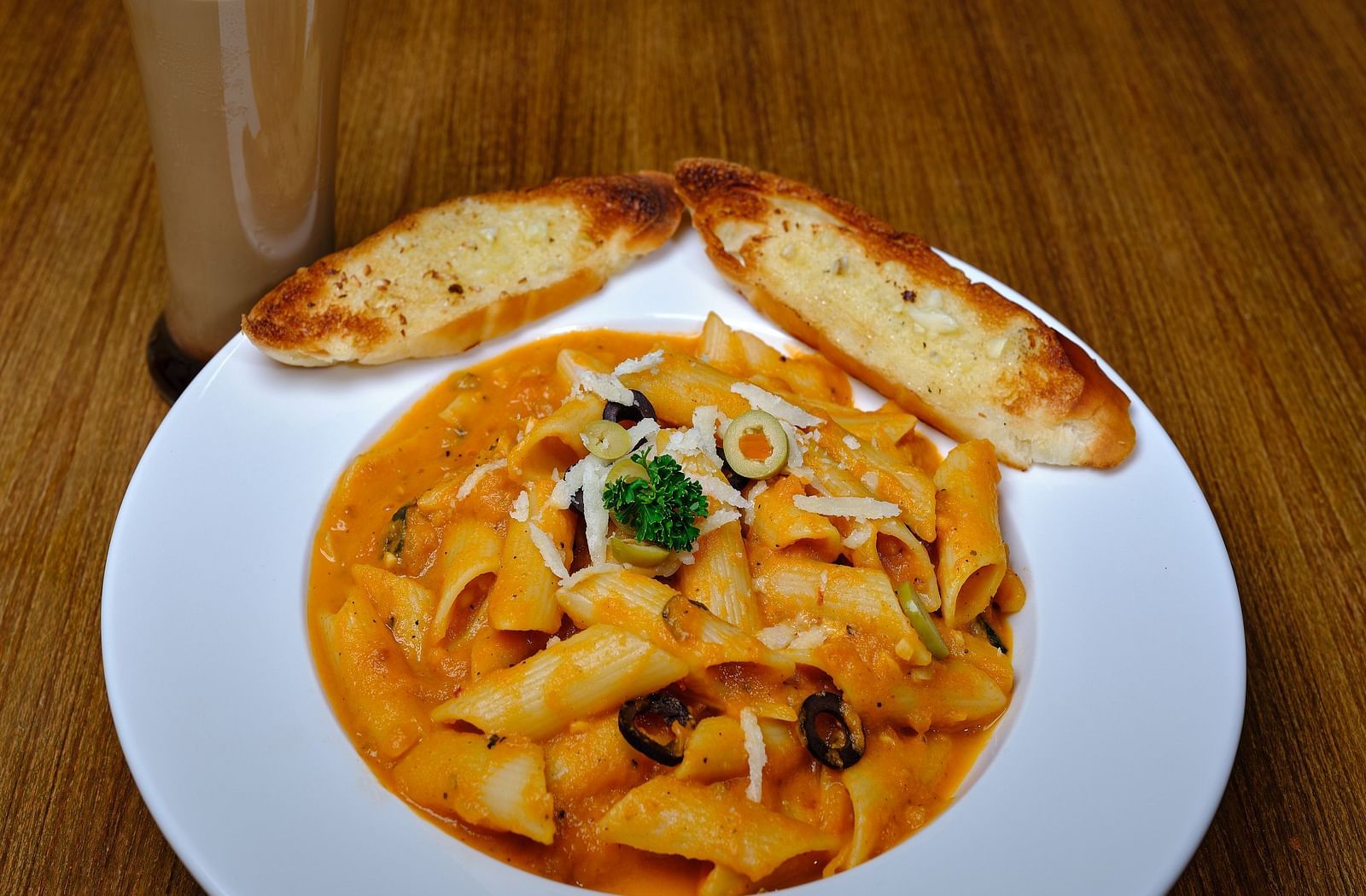 Pasta served in a wide mouth bowl with two pieces of toast and a glass of cold coffee