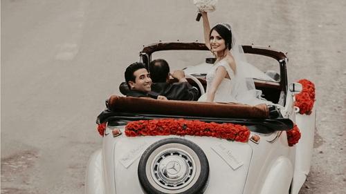 A bride and groom pictured celebrating post wedding on an antique car with wedding decors while a driver is driving the car.