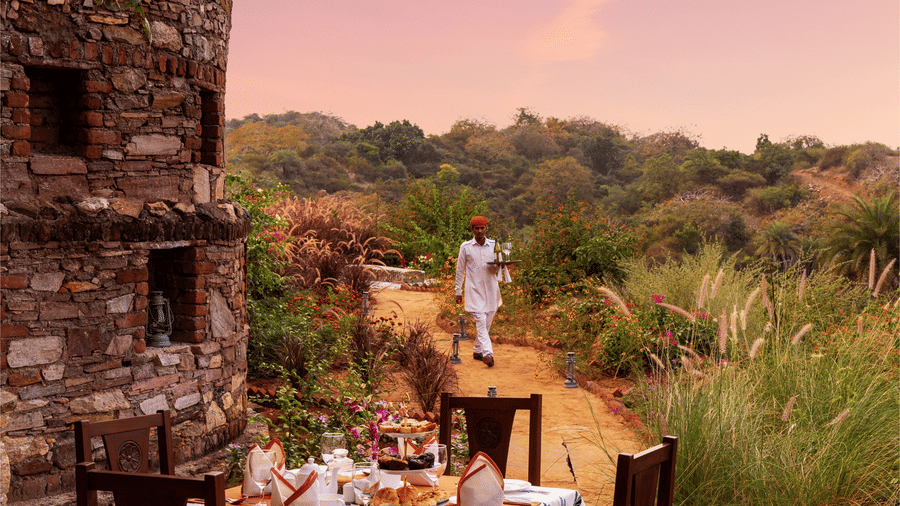 A table for 4 set up next to a stone structure surrounded with greenery and a beautiful sky with pink hues in the background - Chunda Shikar Oudi, Udaipur.