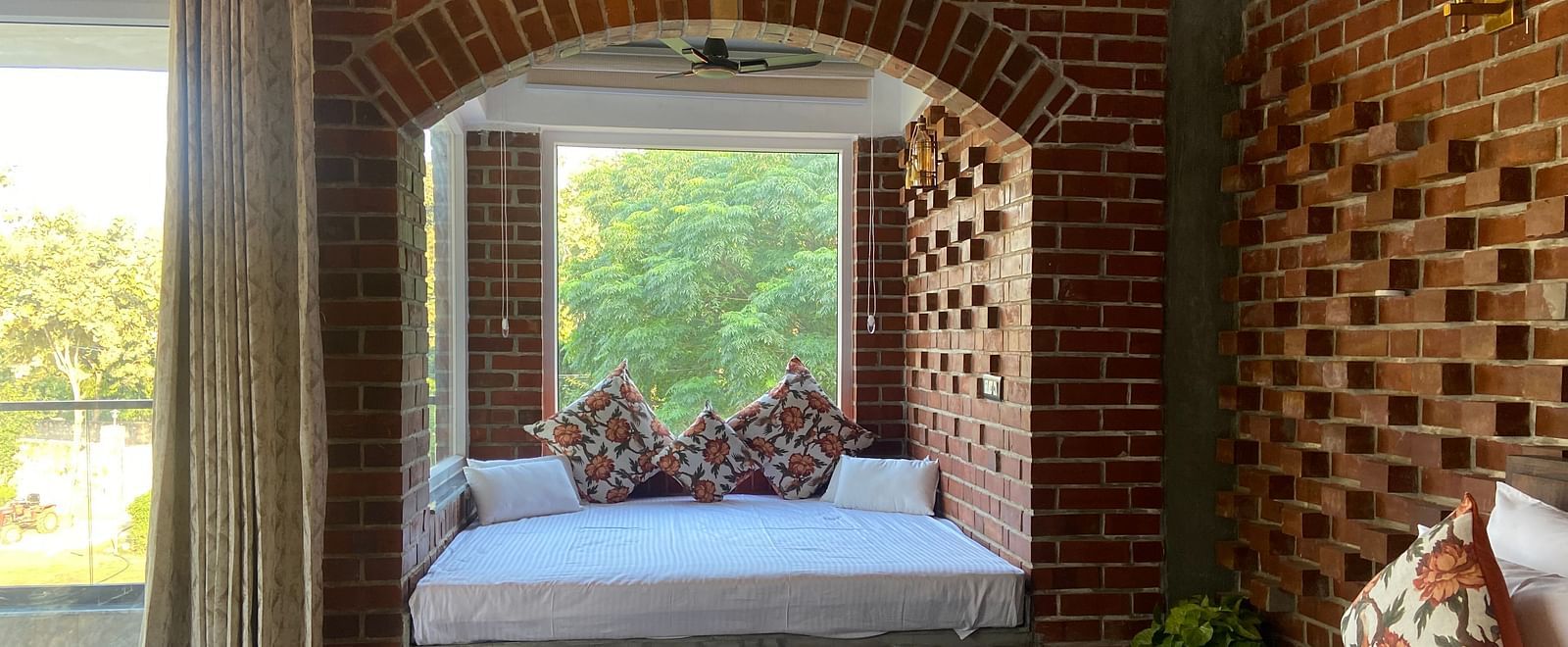 A bedroom at Tisya Farms, Gurgaon featuring a bed near a large arched window, brick walls, and a curtain on one side.
