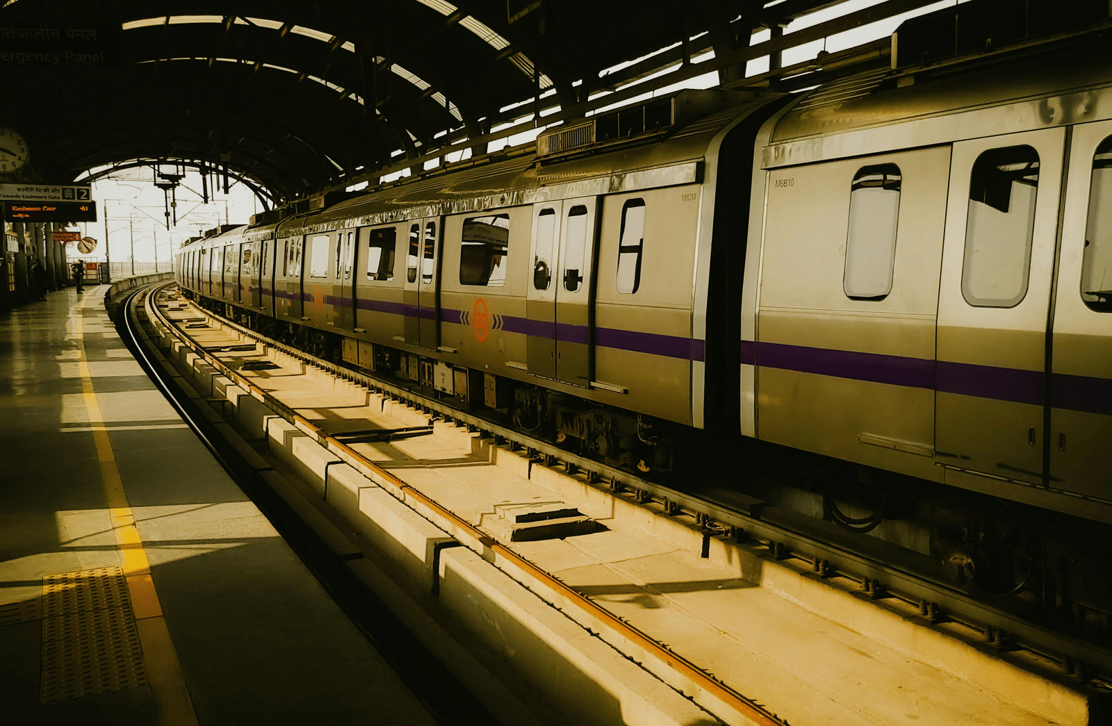 A metro train stationary at an indoor platform station, viewed down the length of the track under a curved roof.