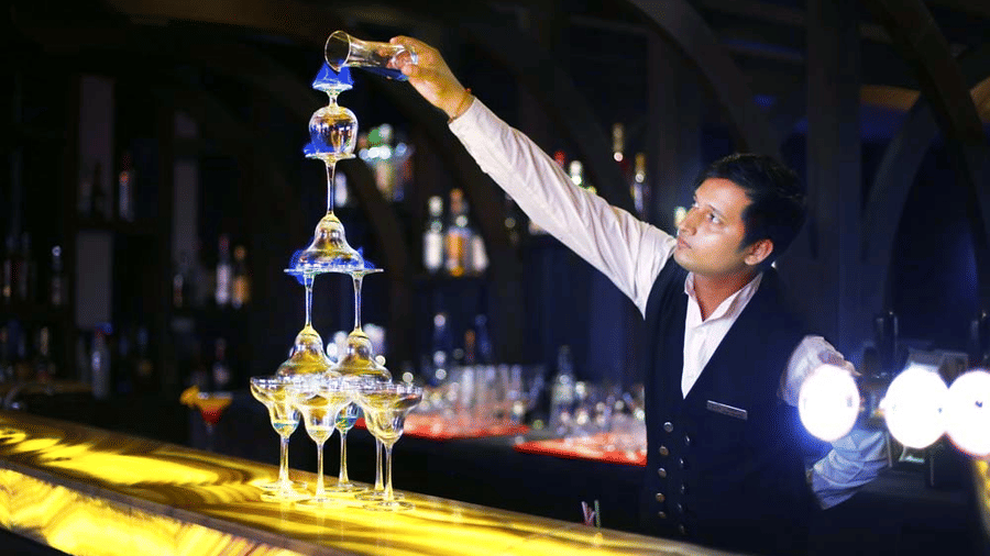 Bartender at The Bristol Hotel, Gurgaon pouring blue liqueur down a tiered champagne glass tower on a backlit bar counter lined with bottles.