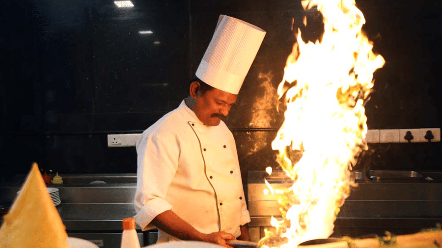 A close-up of a chef in a white toque hat cooking behind a counter as bright orange flames flare high out of a pan.