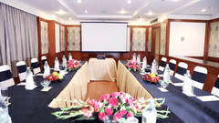 Meeting room at The Bristol Hotel, Gurgaon with tables arranged facing a projector screen, black chair covers, and pink flower arrangements on the tables.