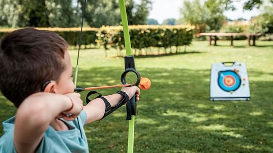  young child, focused and poised, aims a toy bow and arrow at a target in a sunlit garden, practicing archery with concentration.