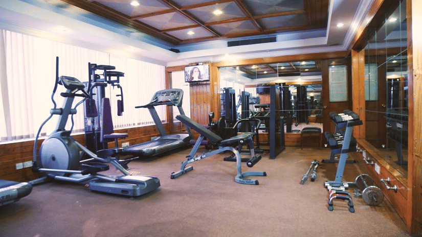 Gym at The Bristol Hotel, Gurgaon with an elliptical trainer, treadmill, weight bench, mirrored wall, wood-panelled ceiling, and a wall-mounted television.