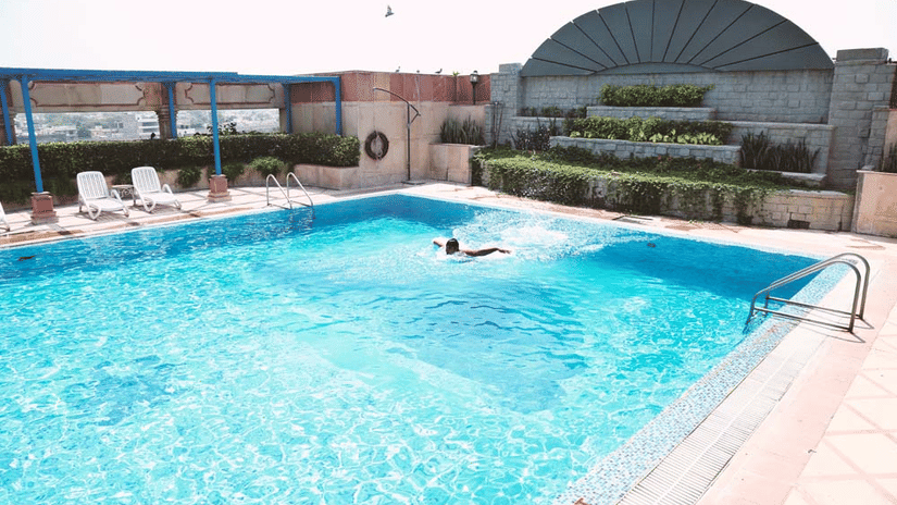 Rooftop swimming pool at The Bristol Hotel, Gurgaon with a person swimming, tiered planters in the background, and a pergola with lounge chairs beside the pool.