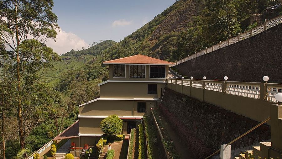 A covered outdoor patio or walkway with a grey metal roof, tiled floor, and a view of trees and a hillside - Abad Copper Castle, Munnar